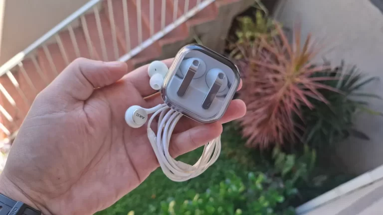 Fotografia em close-up de uma mão segurando um estojo de acrílico transparente. Dentro do estojo está um fone de ouvido bluetooth branco. Fora do estojo, um par de fones de ouvido brancos com fio está enrolado e organizado. O fundo mostra uma planta suculenta desfocada.