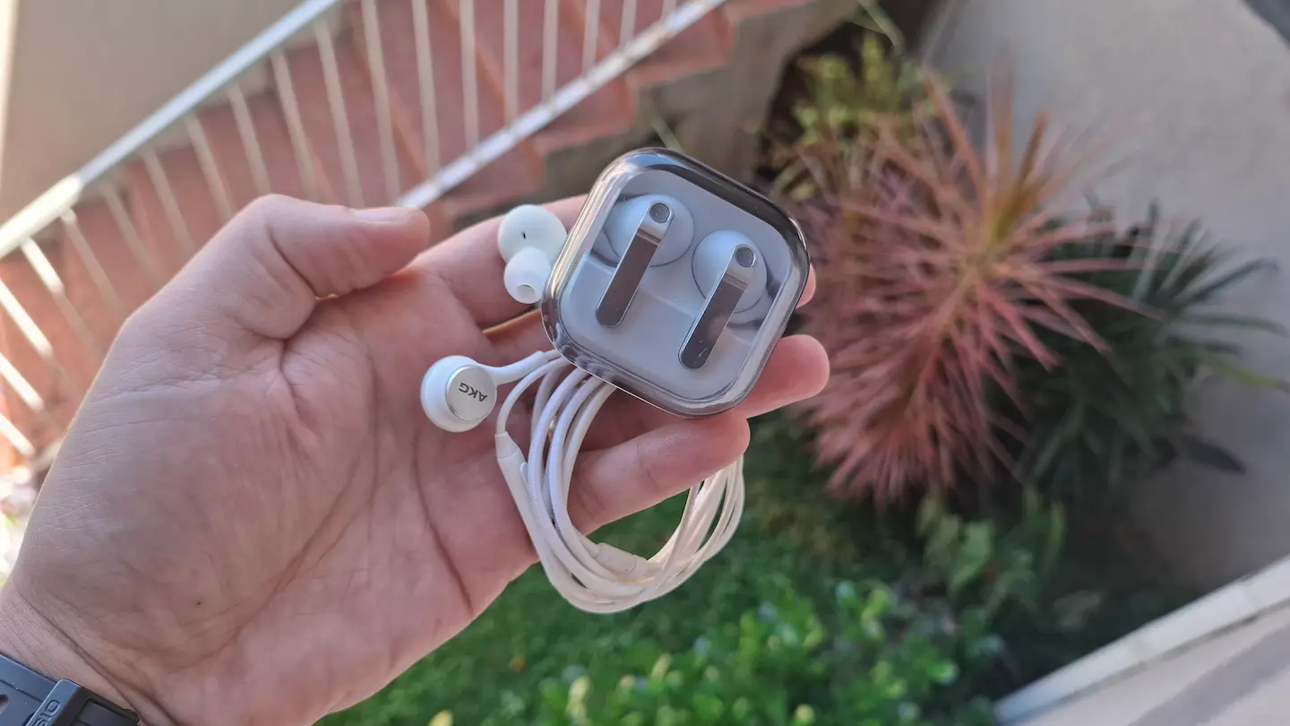 Fotografia em close-up de uma mão segurando um estojo de acrílico transparente. Dentro do estojo está um fone de ouvido bluetooth branco. Fora do estojo, um par de fones de ouvido brancos com fio está enrolado e organizado. O fundo mostra uma planta suculenta desfocada.