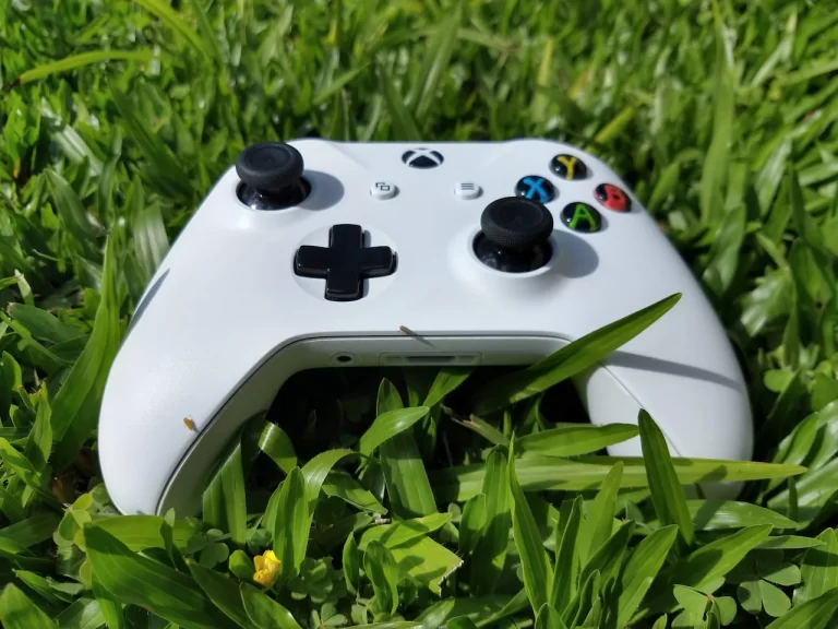 Controle de Xbox One S branco (modelo 1708) em cima da grama verde, destacando o botão Xbox integrado na mesma peça frontal.