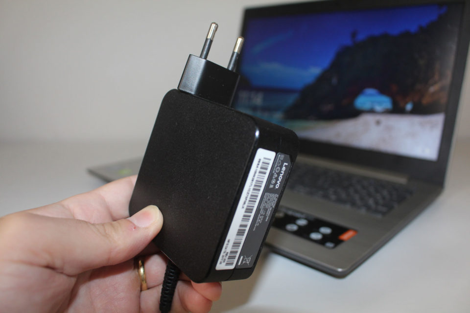Carregador do notebook Lenovo IdeaPad 320 com a fonte integrada à tomada, posicionado em frente ao aparelho.