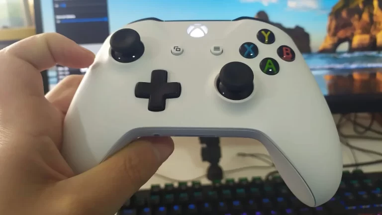 Mão segurando um controle de Xbox One S branco ligado, mostrando o botão power central iluminado e os botões de ação coloridos (A, B, X, Y) em frente a um monitor de PC.