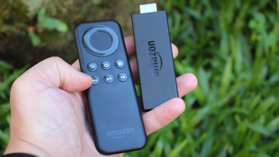 Mão segurando o controle remoto preto e o dispositivo de streaming Amazon Fire TV Stick Basic Edition em primeiro plano, com fundo desfocado de grama verde.