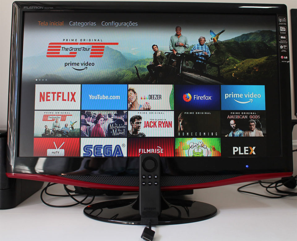 Uma televisão preta de mesa exibindo a interface inicial do Amazon Fire TV Stick com diversos aplicativos de streaming na tela, como Netflix e Prime Video, com o controle remoto posicionado em frente à base da TV.