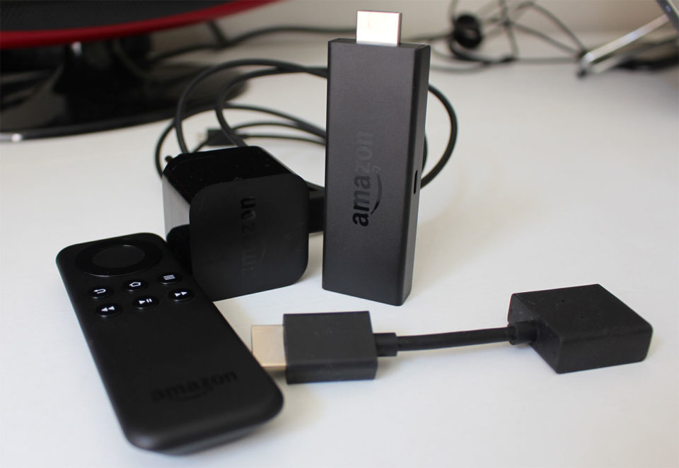 Visão geral dos componentes do Amazon Fire TV Stick sobre a mesa, incluindo o dongle HDMI, o controle remoto preto, o adaptador de energia e cabos conectores.