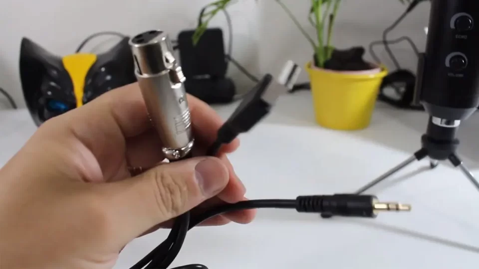 Close de uma mão segurando o cabo híbrido do microfone BM 100FX, mostrando claramente o conector XLR fêmea, a ponta P2 (3.5mm) e o conector USB para alimentação.