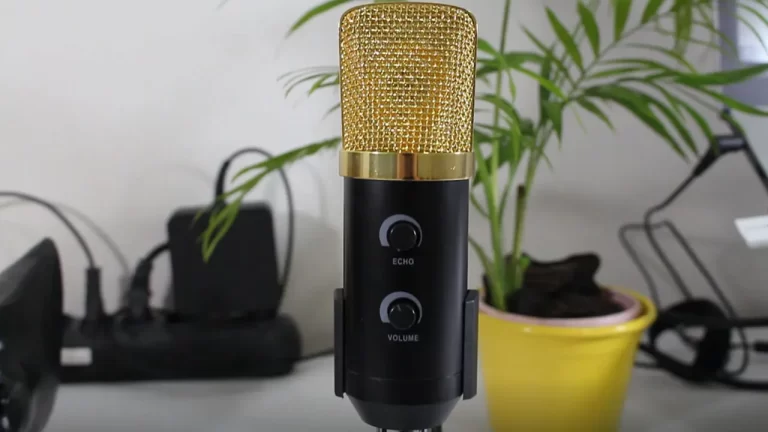 Close do microfone condensador preto com grade dourada BM-100FX, focado nos botões de Echo e Volume, posicionado em um tripé de mesa em um escritório.
