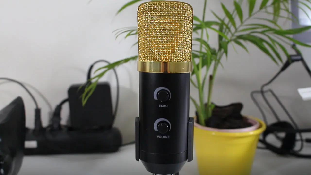 Close do microfone condensador preto com grade dourada BM-100FX, focado nos botões de Echo e Volume, posicionado em um tripé de mesa em um escritório.