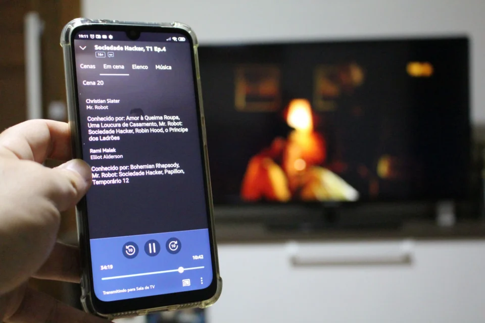 Mão segurando um celular com o aplicativo Amazon Prime Video aberto na tela de reprodução, enquanto transmite um filme para uma televisão ao fundo em ambiente residencial escuro.