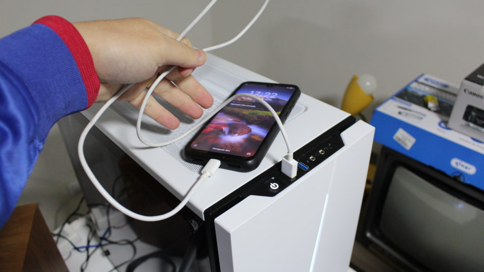 Mão conectando um cabo USB em um smartphone Android posicionado sobre um computador branco para configuração de webcam.