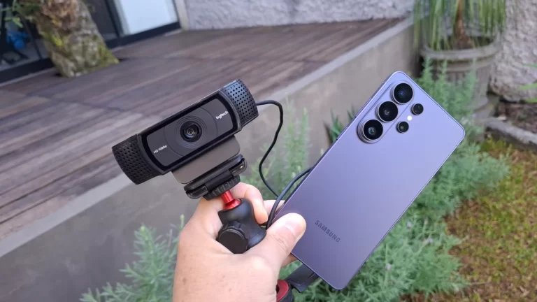 Uma mão segura uma webcam USB comum em um tripé pequeno ao lado de um smartphone Samsung S26 Ultra, comparando os dois dispositivos para uso em videochamadas.