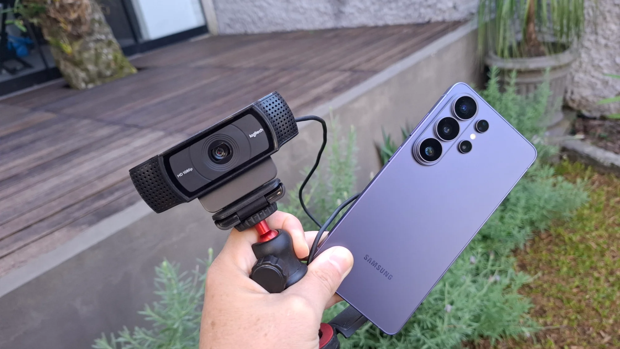 Uma mão segura uma webcam USB comum em um tripé pequeno ao lado de um smartphone Samsung S26 Ultra, comparando os dois dispositivos para uso em videochamadas.