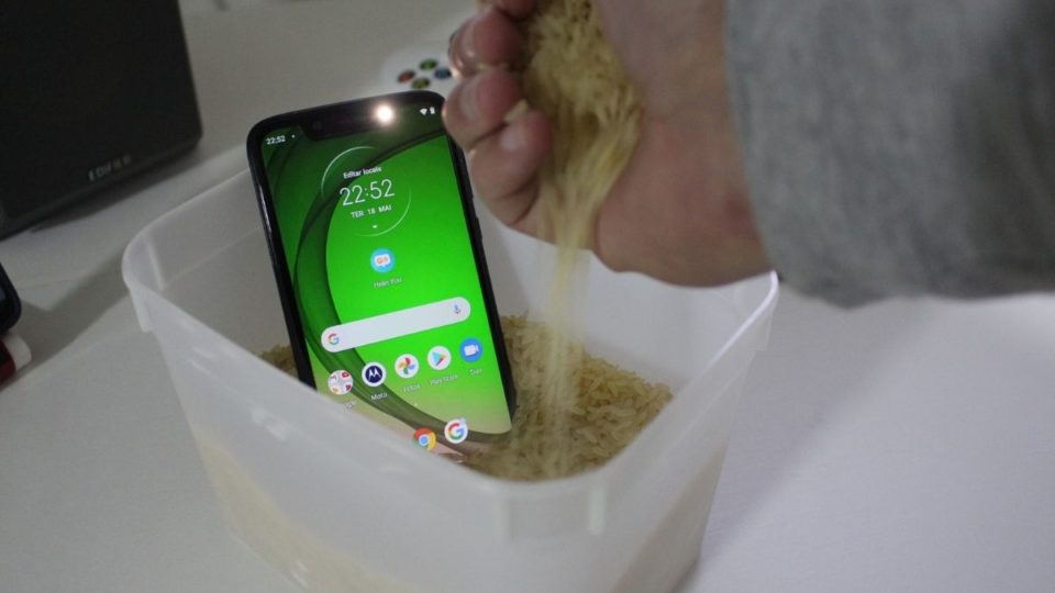 Alguém despejando arroz cru sobre um smartphone molhado dentro de um pote plástico branco.