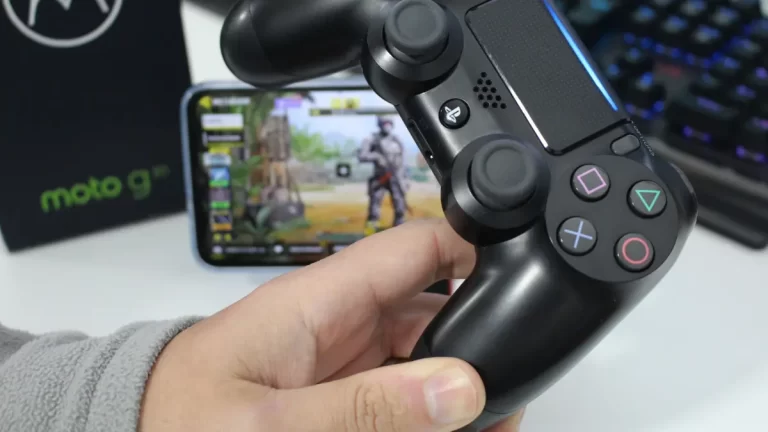 Mão segurando um controle DualShock 4 (PS4) preto ao lado de um smartphone mostrando uma interface de jogo.
