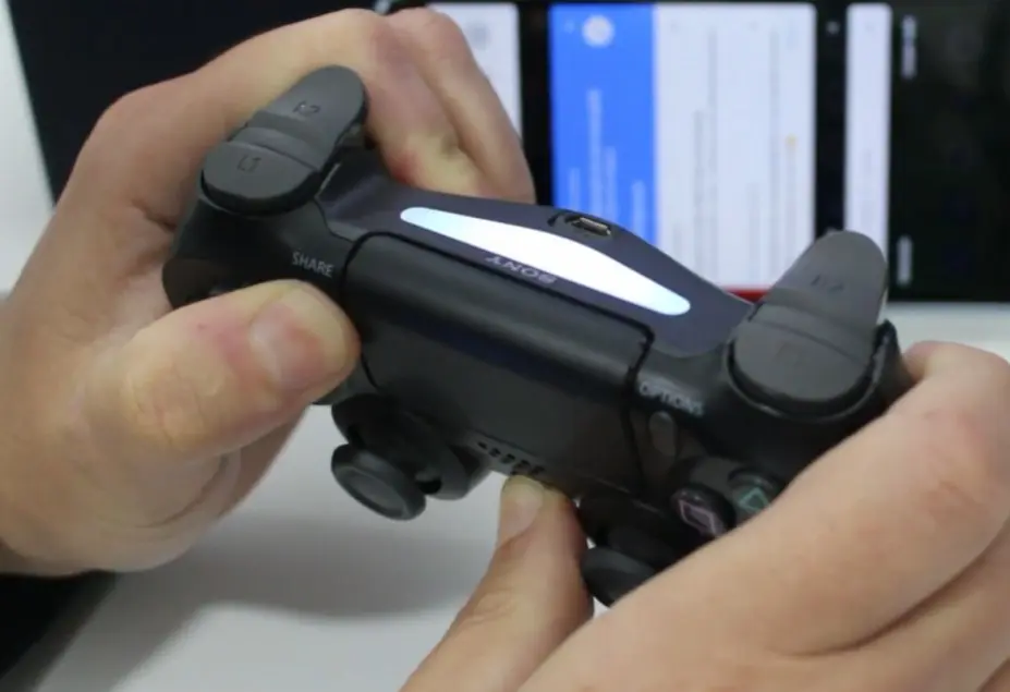 Close em uma mão segurando o controle de PS4 preto e mostrando a barra de luz ligada em branco, indicando modo de pareamento.