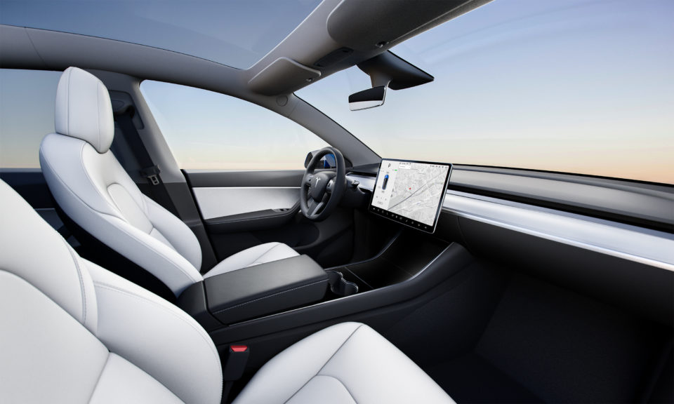 Visão interna do Tesla Model Y destacando os bancos de couro branco, o teto solar panorâmico de vidro e o painel minimalista com a tela central de 15 polegadas.