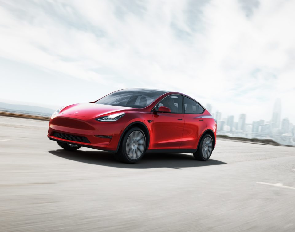 Tesla Model Y na cor vermelha em movimento em uma estrada, visto de ângulo frontal diagonal. O veículo destaca o design aerodinâmico e as rodas esportivas escuras sob um céu nublado.