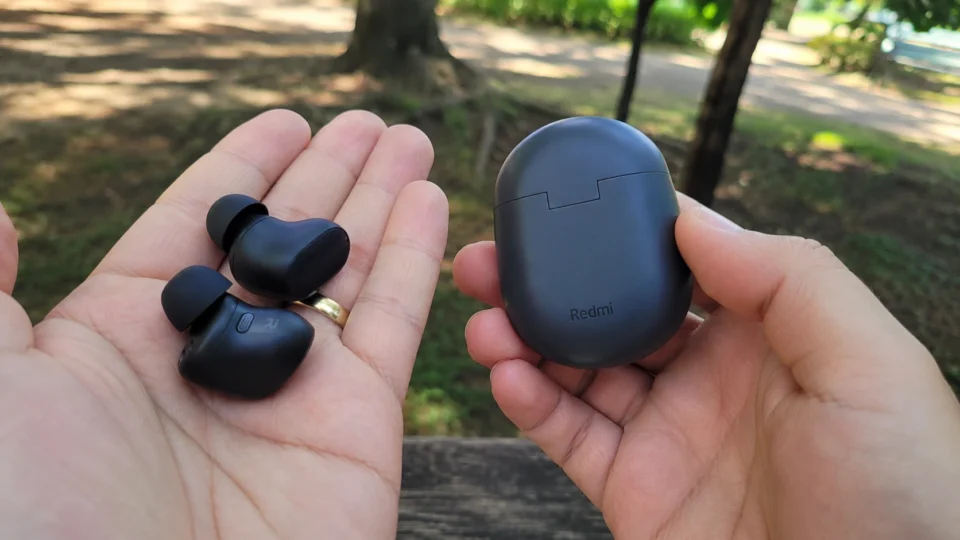Mão esquerda segurando os dois fones de ouvido Bluetooth pretos Redmi Buds 3 Pro abertos na palma, enquanto a mão direita segura o estojo de carregamento oval cinza-escuro com fundo desfocado de árvores ao ar livre.