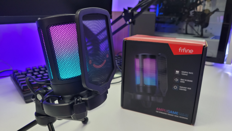 Microfone Fifine AmpliGame A6V com iluminação RGB ao lado de sua caixa original em um setup gamer.