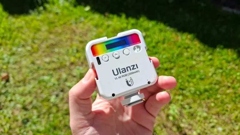 Mão segurando o mini refletor LED Ulanzi VL49 RGB branco em ambiente externo com luz solar, destacando a paleta de cores HSI no topo e sua construção compacta.