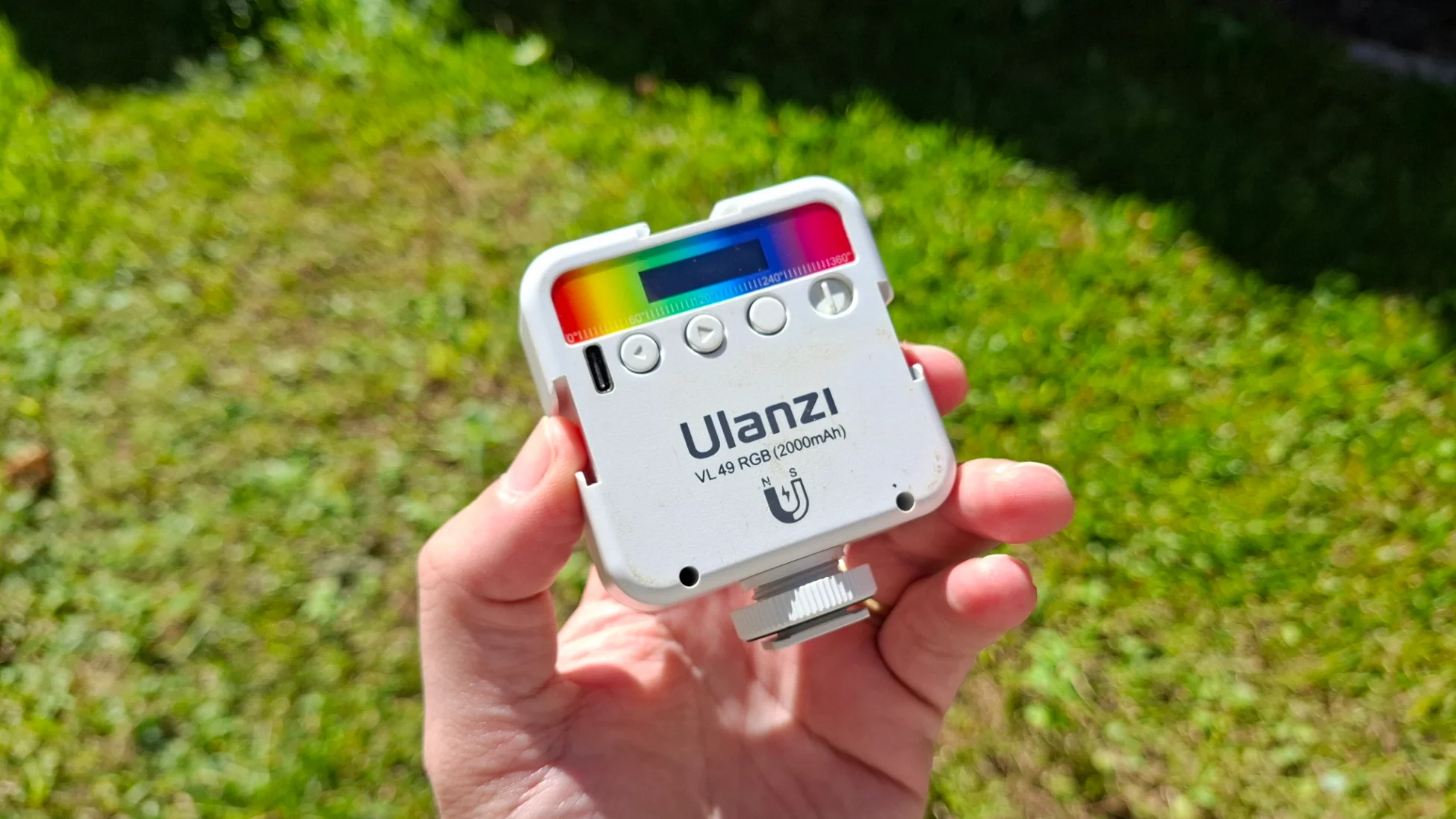 Mão segurando o mini refletor LED Ulanzi VL49 RGB branco em ambiente externo com luz solar, destacando a paleta de cores HSI no topo e sua construção compacta.