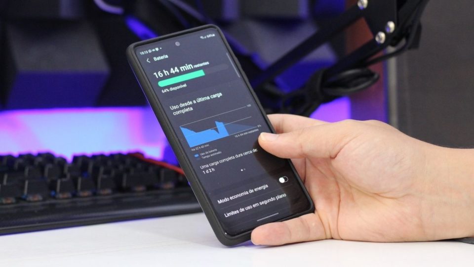 Mão segurando um smartphone Samsung Galaxy S20 FE exibindo gráficos de uso de bateria em uma tela com fundo escuro, próximo a um teclado gamer.