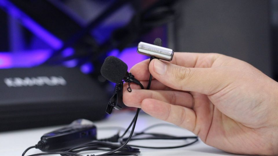 Detalhe de um microfone de lapela preto com clipe de fixação sendo segurado próximo a um receptor sem fio.
