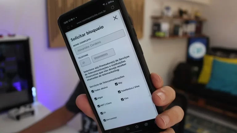 Pessoa segurando um smartphone que mostra a tela de cadastro do site Não Me Perturbe para bloqueio de ligações, com um escritório ao fundo.