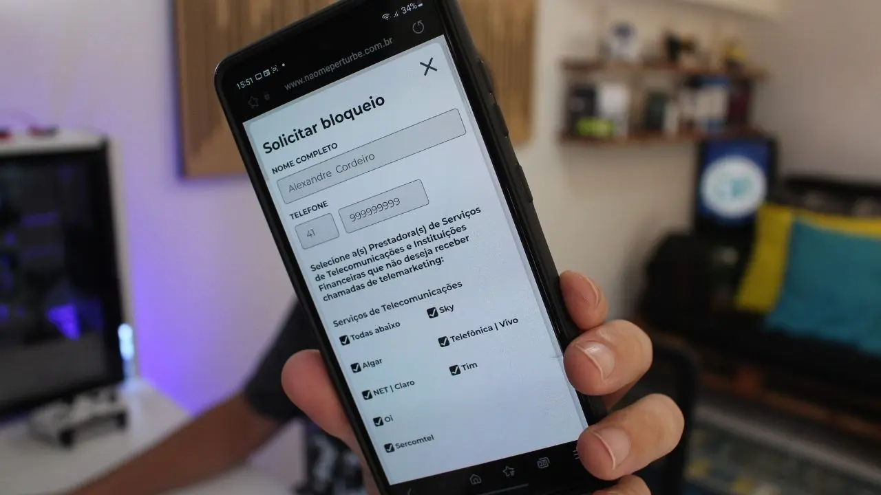 Pessoa segurando um smartphone que mostra a tela de cadastro do site Não Me Perturbe para bloqueio de ligações, com um escritório ao fundo.
