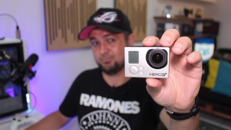 Close-up das mãos de um técnico segurando uma câmera GoPro Hero antiga, preparando-a para ser usada como webcam de alta qualidade em um Home Office com iluminação RGB.