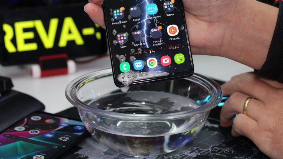 Mão segurando um smartphone parcialmente submerso em uma tigela de vidro com água limpa, ilustrando o momento de um acidente com líquido.