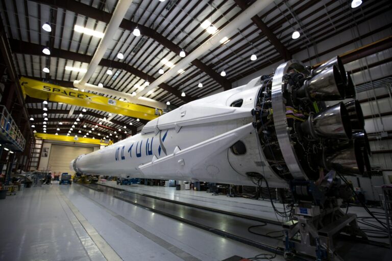 Fotografia do interior de um galpão industrial da SpaceX mostrando um foguete branco de grande porte em posição horizontal sobre trilhos. É possível ver detalhes dos motores na parte traseira.