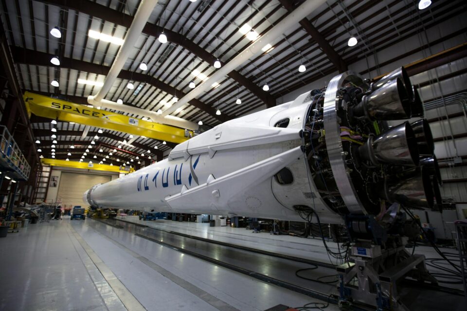 Fotografia do interior de um galpão industrial da SpaceX mostrando um foguete branco de grande porte em posição horizontal sobre trilhos. É possível ver detalhes dos motores na parte traseira.