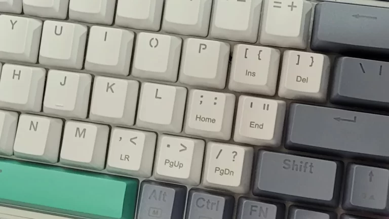 Teclado mecânico compacto com layout internacional (ANSI) em tons de cinza e verde sobre uma mesa.