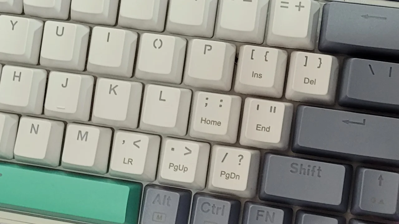 Teclado mecânico compacto com layout internacional (ANSI) em tons de cinza e verde sobre uma mesa.