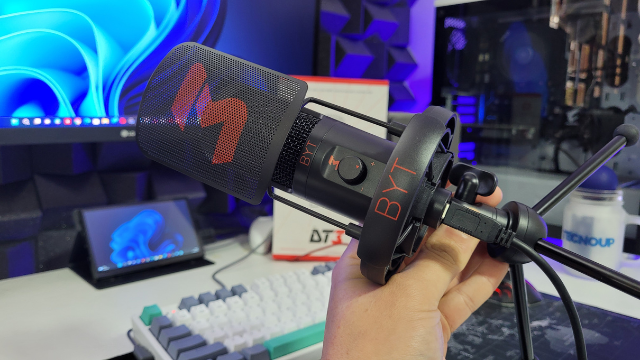 Mão segurando o microfone DT3 BYT preto em frente a um setup gamer com monitor e teclado mecânico ao fundo.