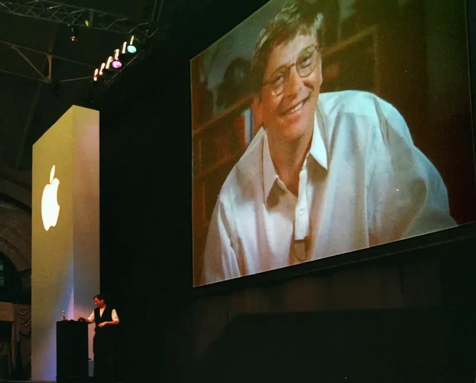 Fotografia histórica da Macworld Expo 1997 mostrando Steve Jobs no palco à esquerda e uma imagem gigante de Bill Gates sorrindo no telão ao fundo.