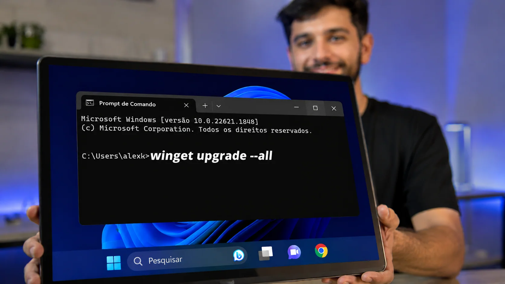 Rapaz segurando uma tela que exibe o Prompt de Comando do Windows com o comando winget upgrade all digitado na tela preta.