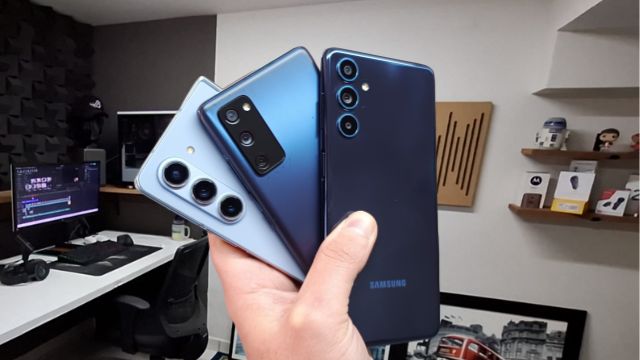Três smartphones Samsung alinhados na mão de Alexandre Cordeiro: o topo de linha dobrável Z Fold 5, o equilibrado S20 FE e o intermediário M55. As câmeras traseiras e os designs distintos de cada modelo são visíveis sobre um fundo desfocado.