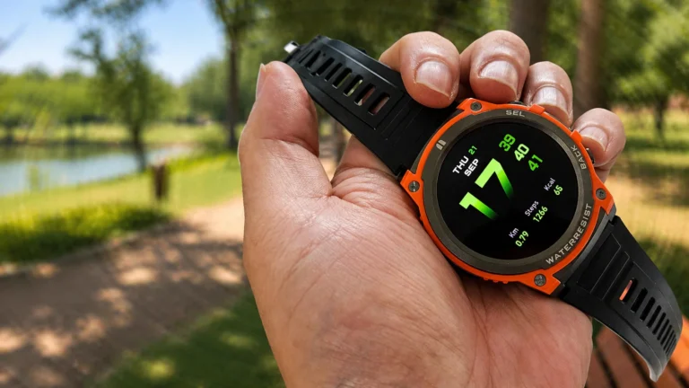 Mão segurando o smartwatch MASX Aurora One com aro laranja e tela ligada, em um parque ensolarado com árvores ao fundo.