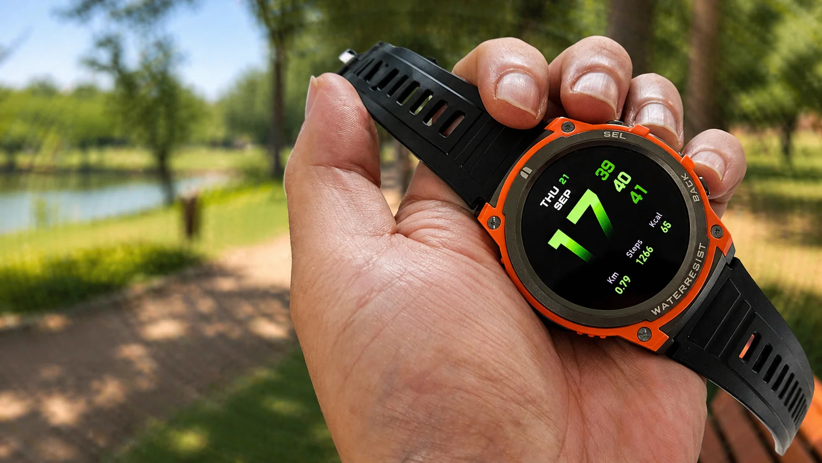 Mão segurando o smartwatch MASX Aurora One com aro laranja e tela ligada, em um parque ensolarado com árvores ao fundo.
