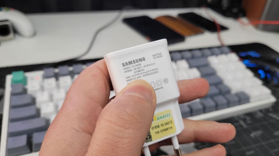 Mão segurando um carregador de parede original Samsung branco modelo EP-TA200B em um ambiente de escritório com teclado ao fundo.