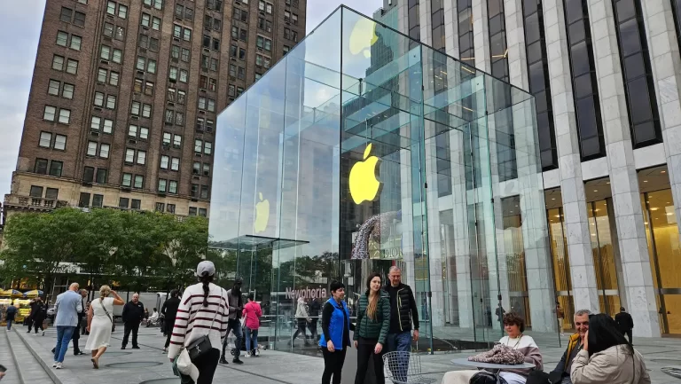O famoso cubo de vidro da Apple Store na Quinta Avenida em Nova York, com pessoas caminhando ao redor e prédios históricos ao fundo.