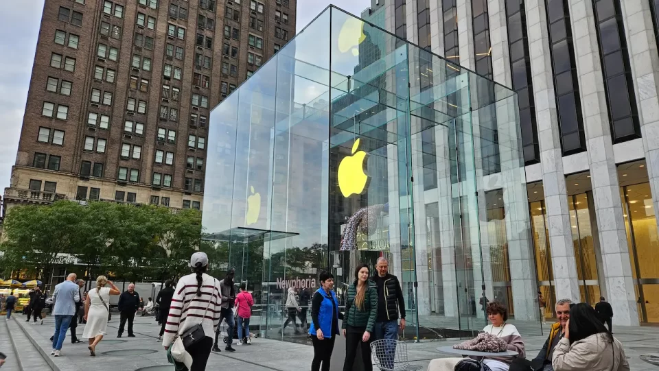 O famoso cubo de vidro da Apple Store na Quinta Avenida em Nova York, com pessoas caminhando ao redor e prédios históricos ao fundo.