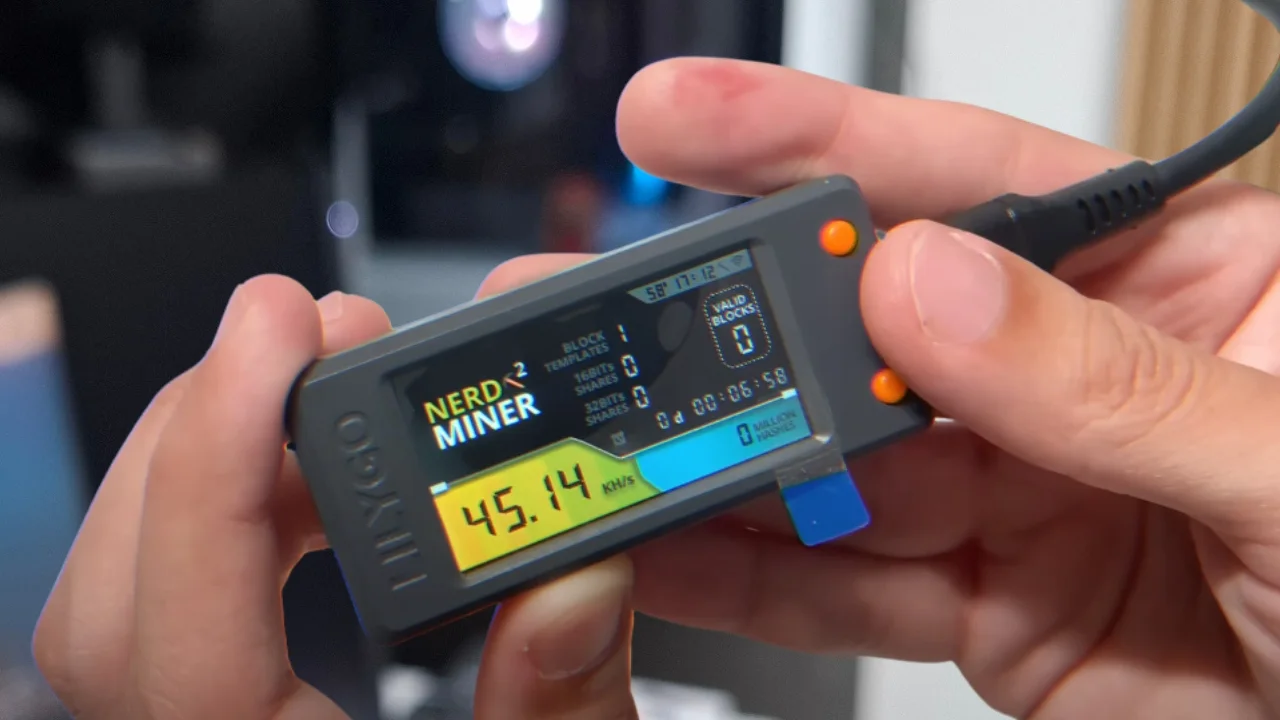 Foto de uma mão segurando um pequeno dispositivo ESP32 com tela LCD ligada, exibindo informações gráficas do sistema de mineração NerdMiner.