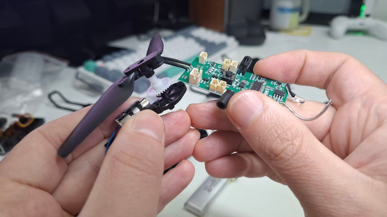 Processo de montagem de um mini drone de brinquedo, mostrando as mãos do Alexandre conectando a placa controladora e os motores.