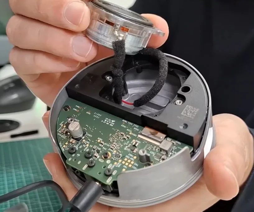 Close das mãos de um técnico removendo o alto-falante de uma Amazon Echo Dot desmontada, revelando a placa eletrônica e os componentes internos.