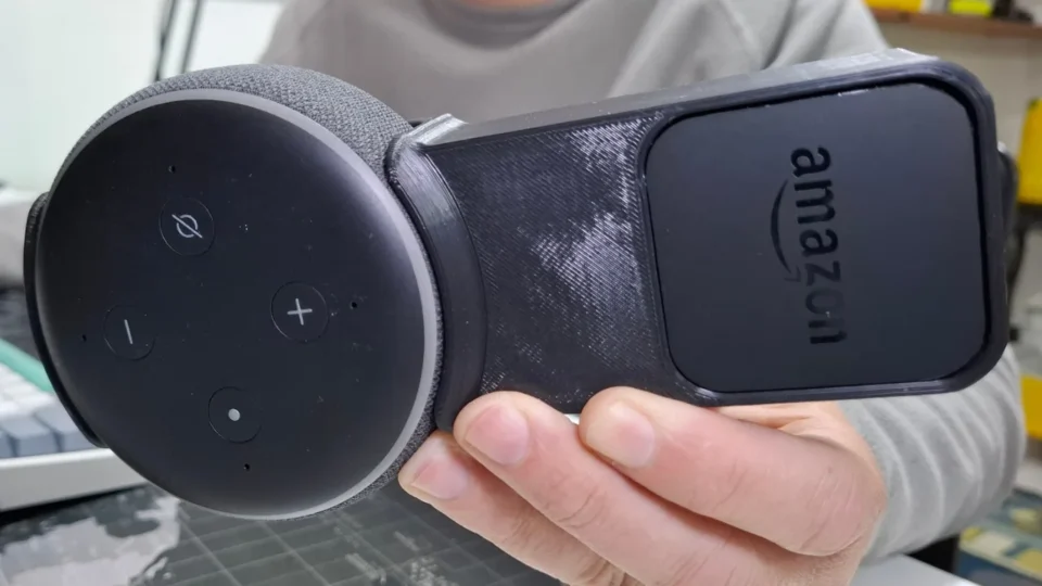 Visão detalhada de uma Alexa Echo Dot 3 instalada em um suporte de tomada preto, mostrando o encaixe perfeito e a ausência de fios aparentes.