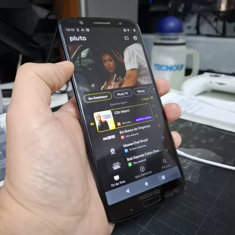 Smartphone exibindo o aplicativo da Pluto TV com a lista de canais ao vivo e interface em modo escuro.