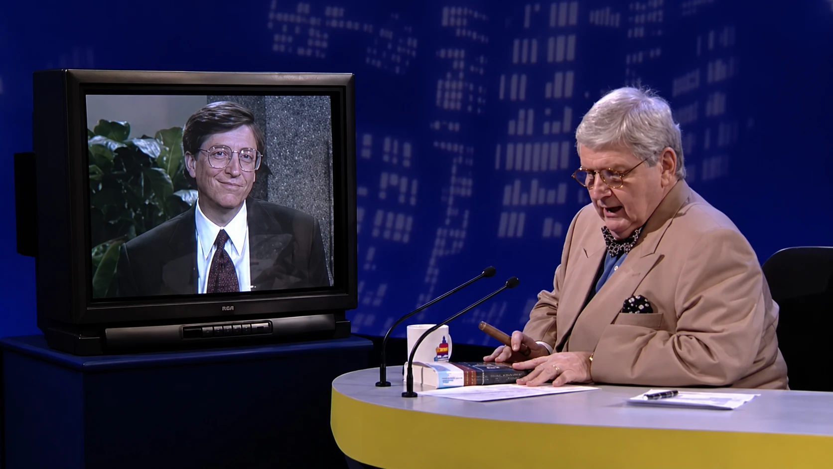 O apresentador Jô Soares sentado em sua mesa de entrevista olhando para baixo enquanto faz anotações, ao lado de um monitor de televisão antigo que exibe Bill Gates sorrindo durante a transmissão via satélite em 1995.