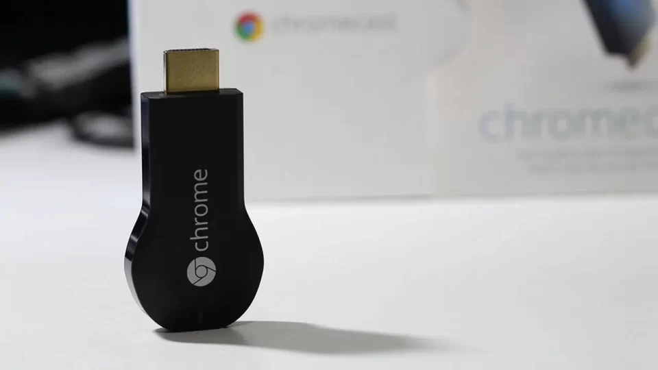 Aparelho Google Chromecast de primeira geração, modelo na cor preta lançado em 2013, deitado sobre uma mesa.